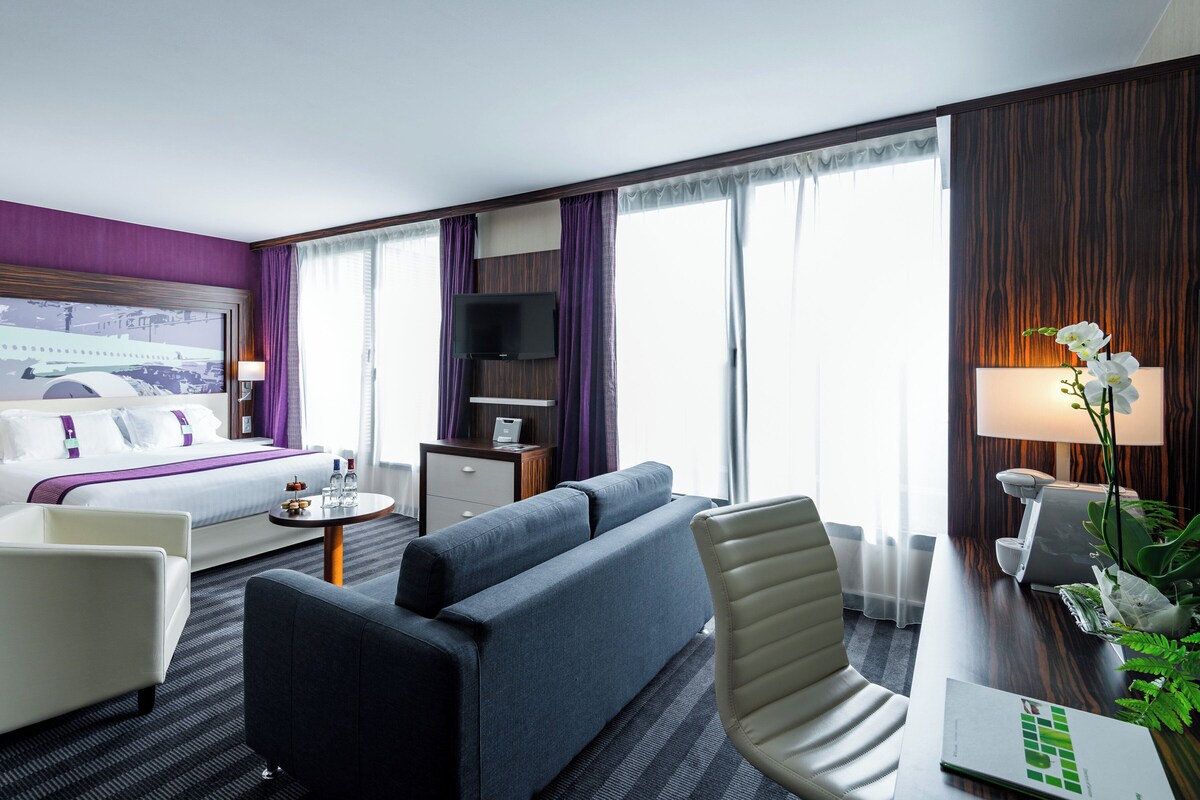 A modern hotel room with a bed, sofa, armchair, desk, and a large window with curtains.