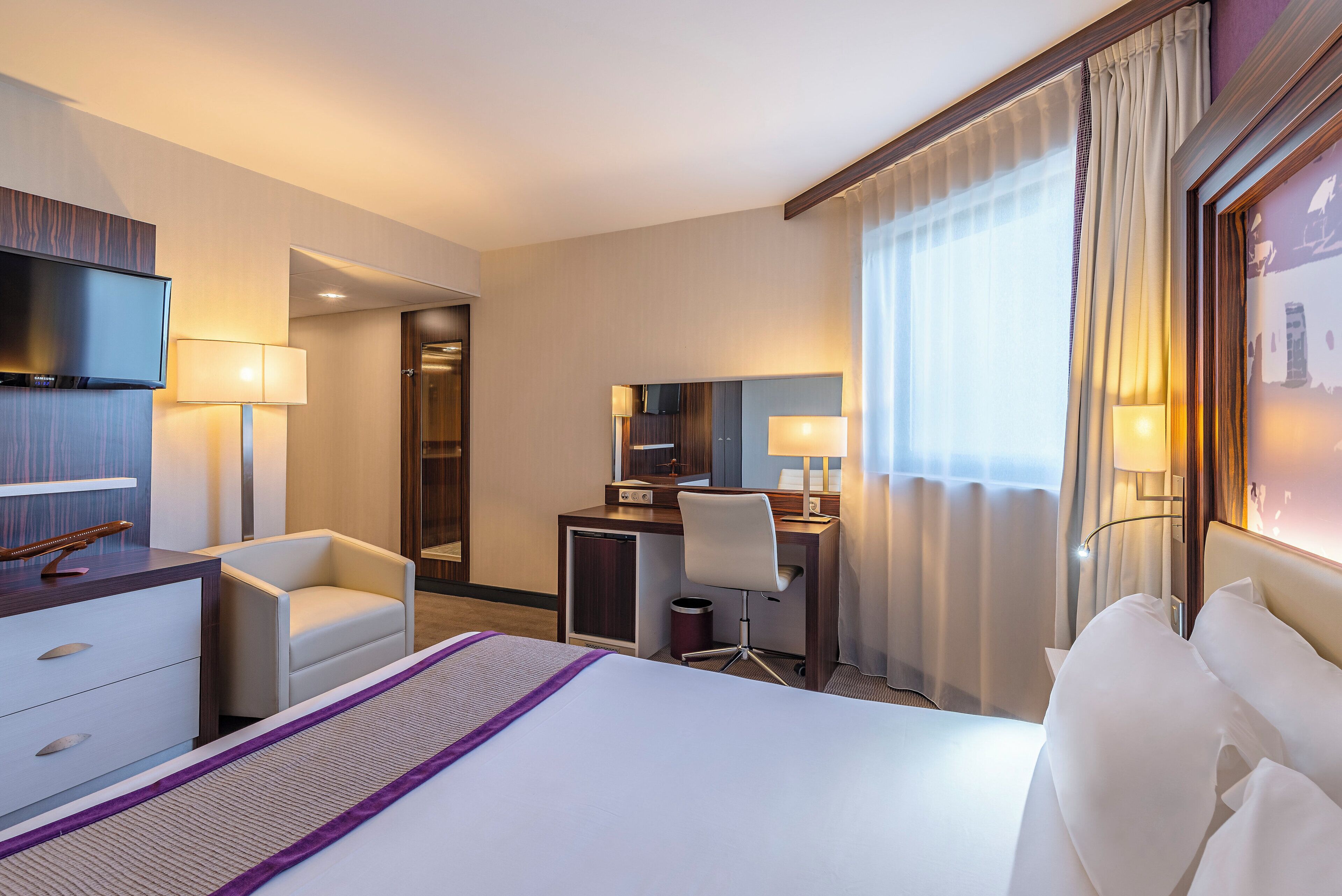 Une chambre d’hôtel comprenant un lit, un bureau avec une chaise, une télévision et une grande fenêtre avec des rideaux.