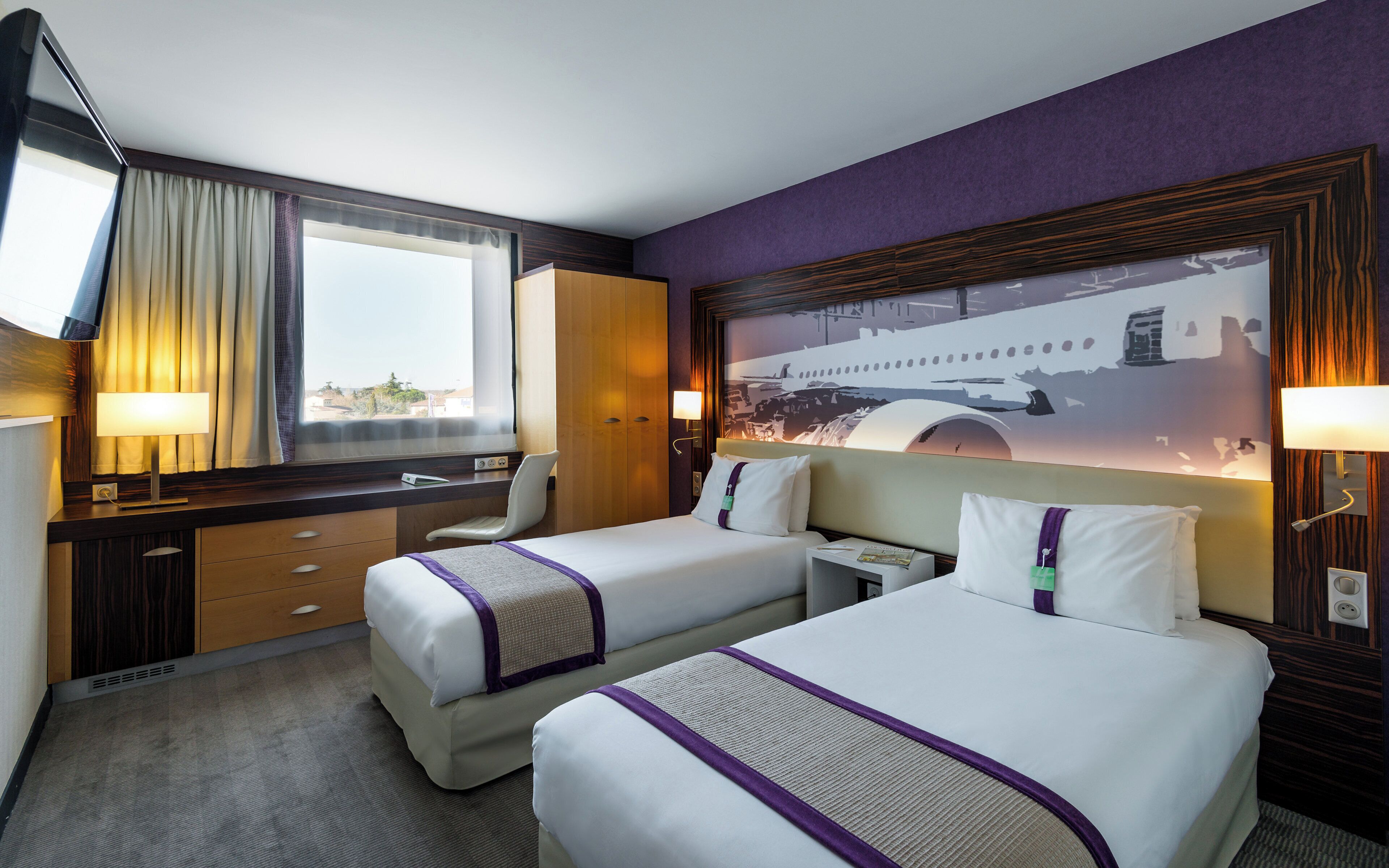 A hotel room with two beds, a desk, a chair, a window with curtains, and a wall-mounted lamp.