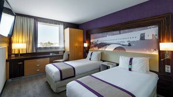 A hotel room with two beds, a desk, a chair, a window with curtains, and a wall-mounted lamp.