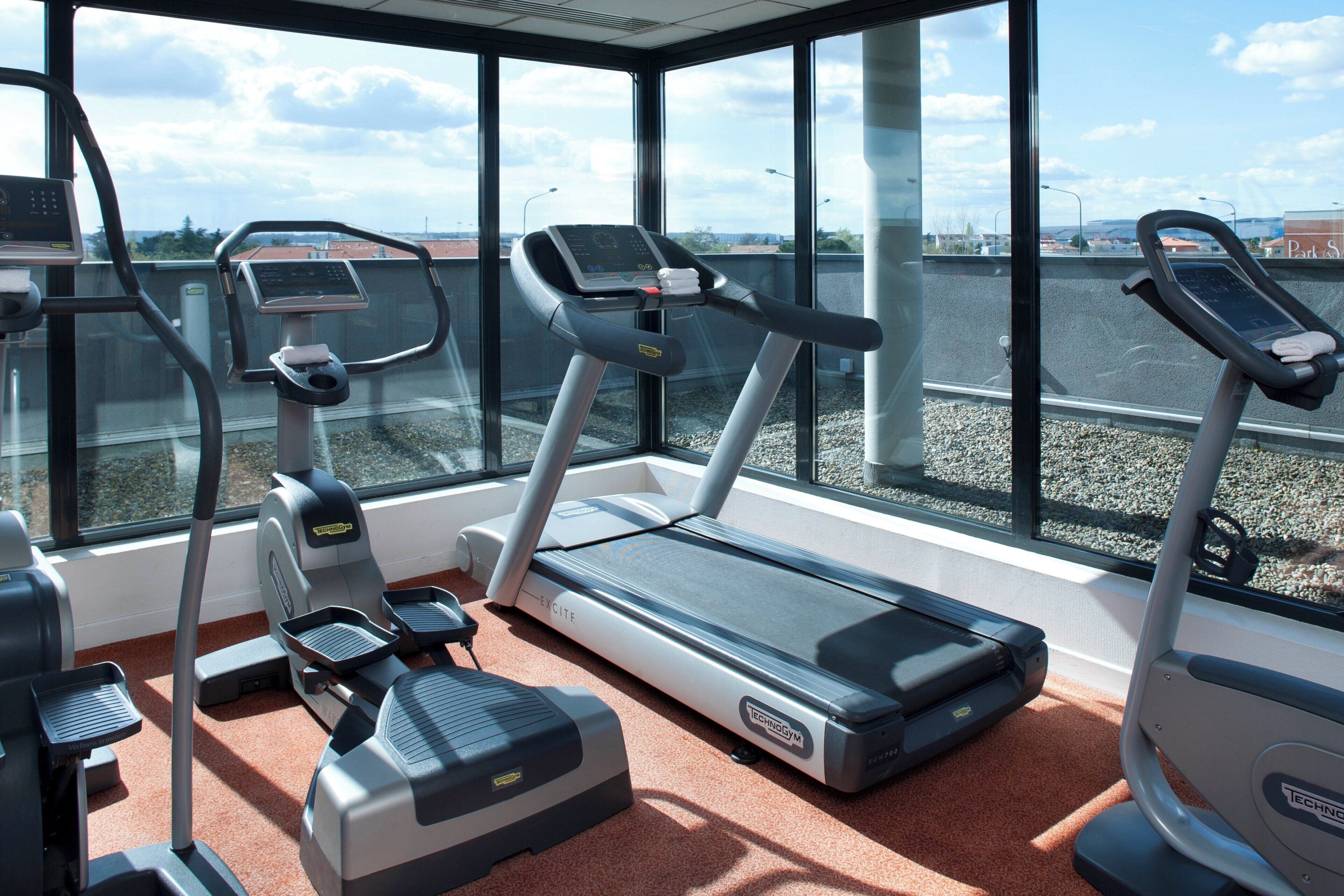 Une salle de sport équipée de tapis de course, d’elliptiques et d’un vélo d’appartement, avec de grandes fenêtres et un sol recouvert d’un tapis rouge.