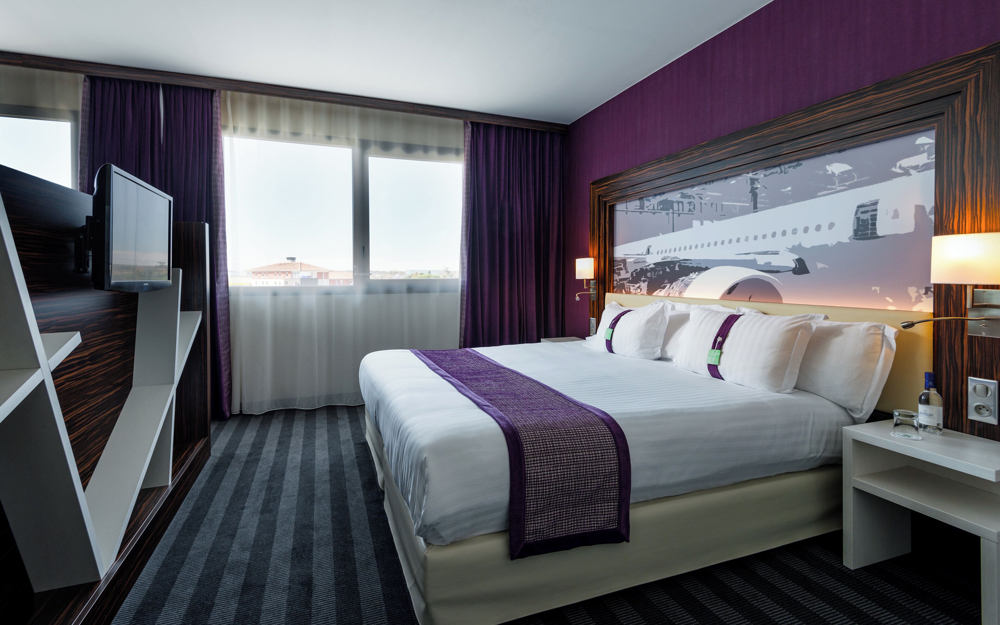 A hotel room with a bed, a TV, a window with curtains, a nightstand, and a wall with a picture.