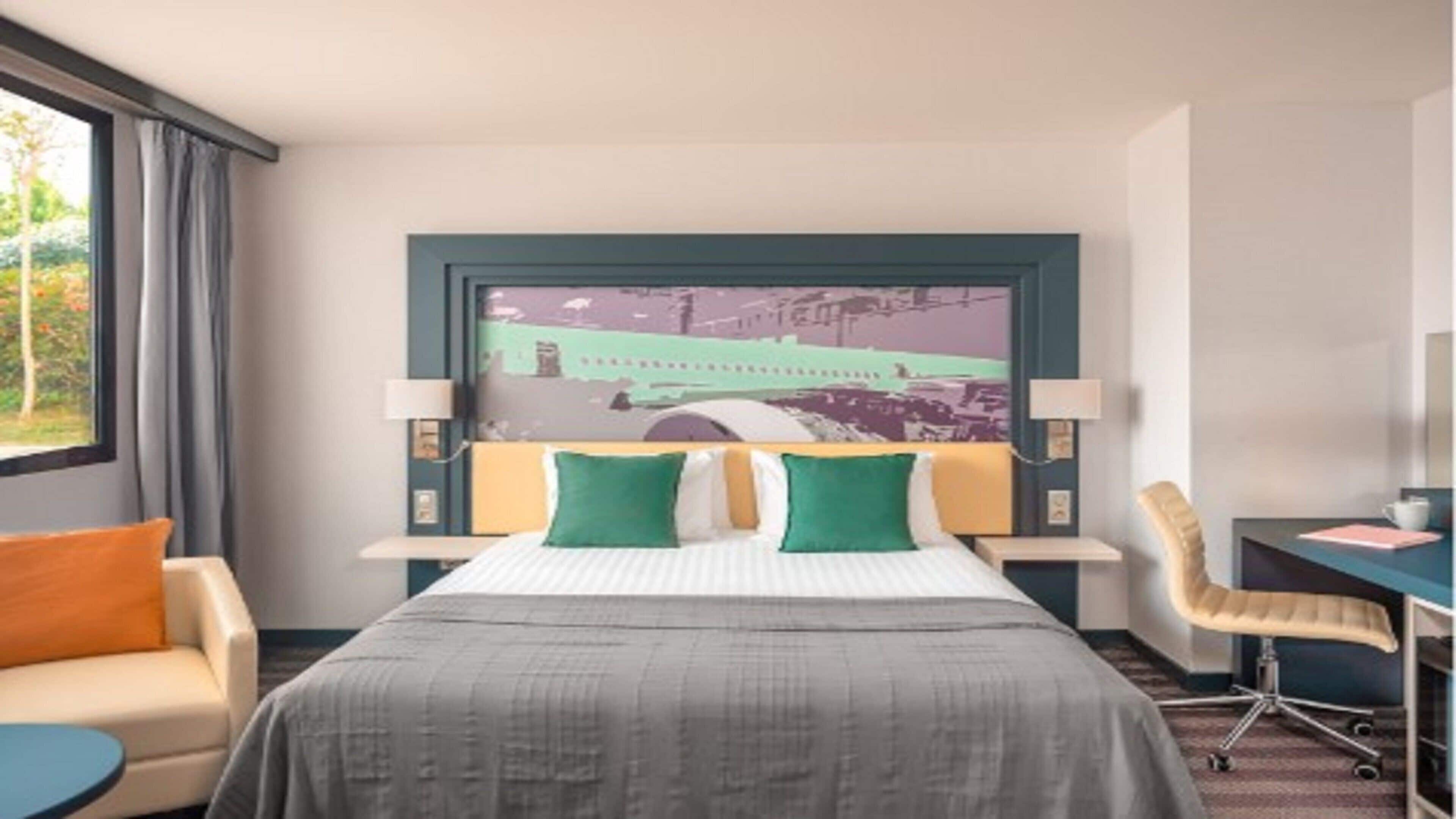 Habitación de hotel con cama, dos almohadas, escritorio, silla, ventana con cortinas y una obra de arte colgada en la pared.
