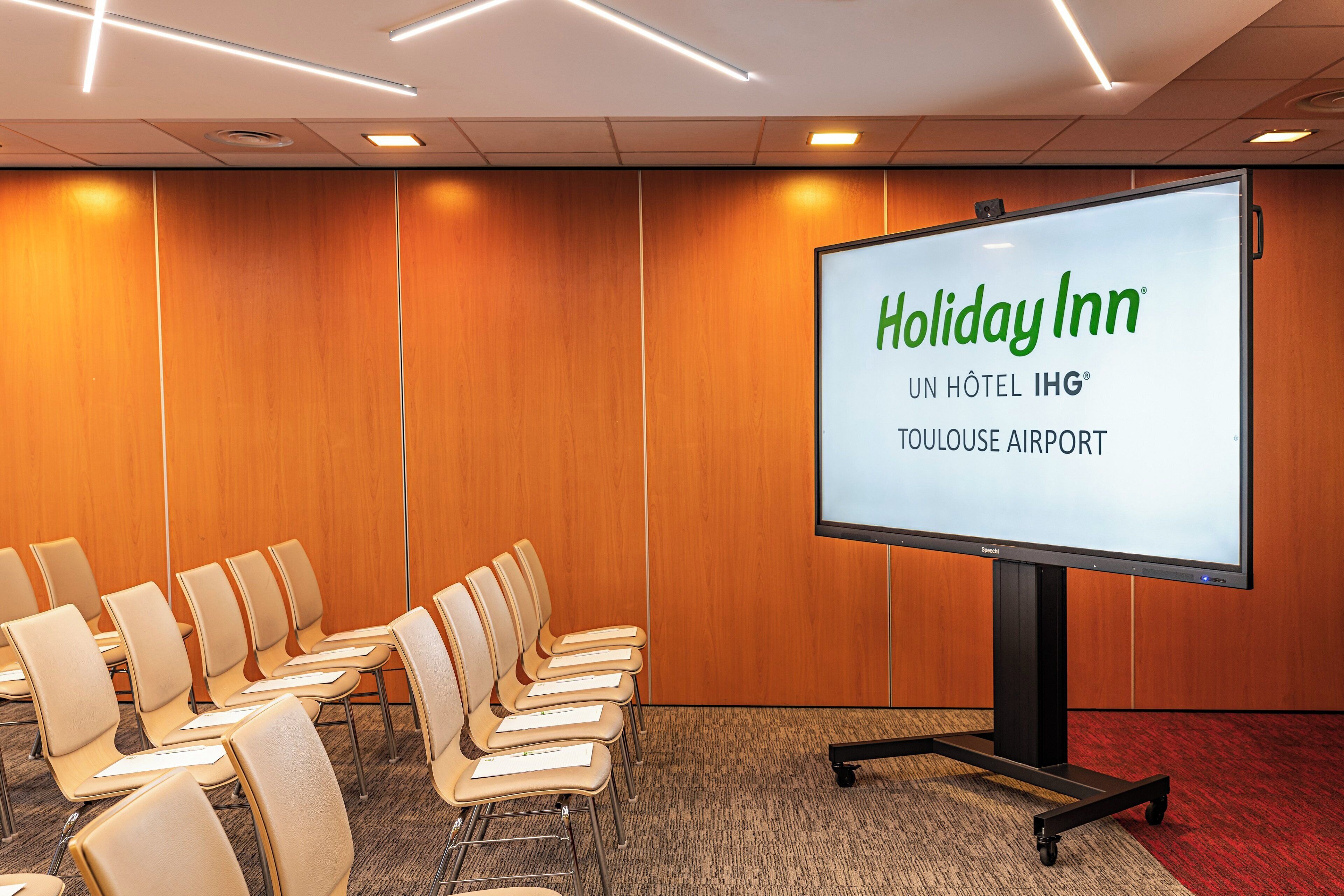 Un salón de conferencias con una pantalla grande que muestra 'Holiday Inn Toulouse Airport' y varias filas de sillas.