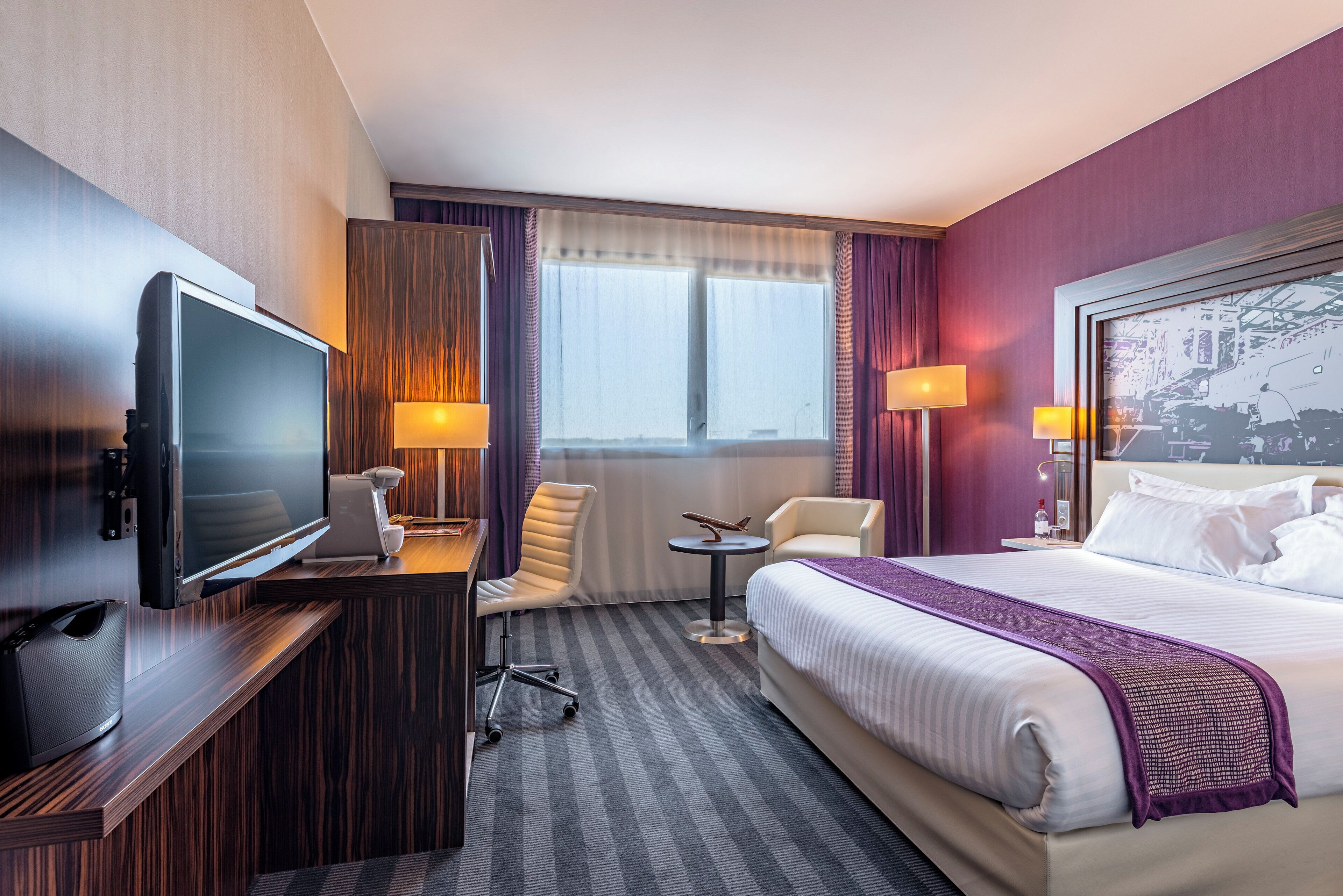 A hotel room with a bed, a desk with a television, a chair, a small table, and a window with curtains.