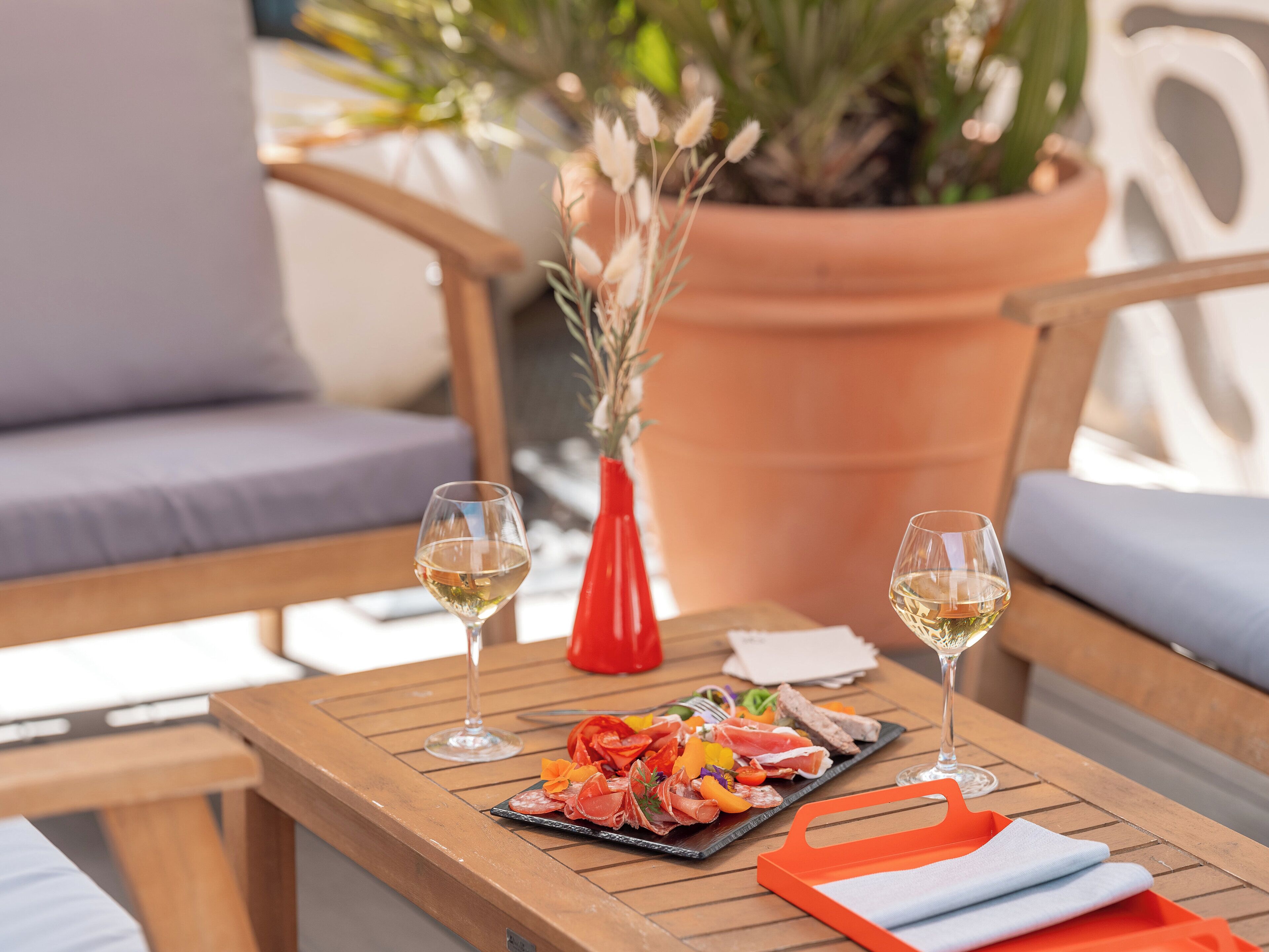 A wooden table with a charcuterie board, two glasses of white wine, and a red vase with dried plants.