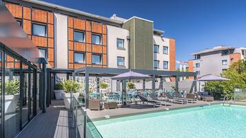 A rooftop pool area with lounge chairs and umbrellas, surrounded by modern buildings with large windows and a clear blue sky.