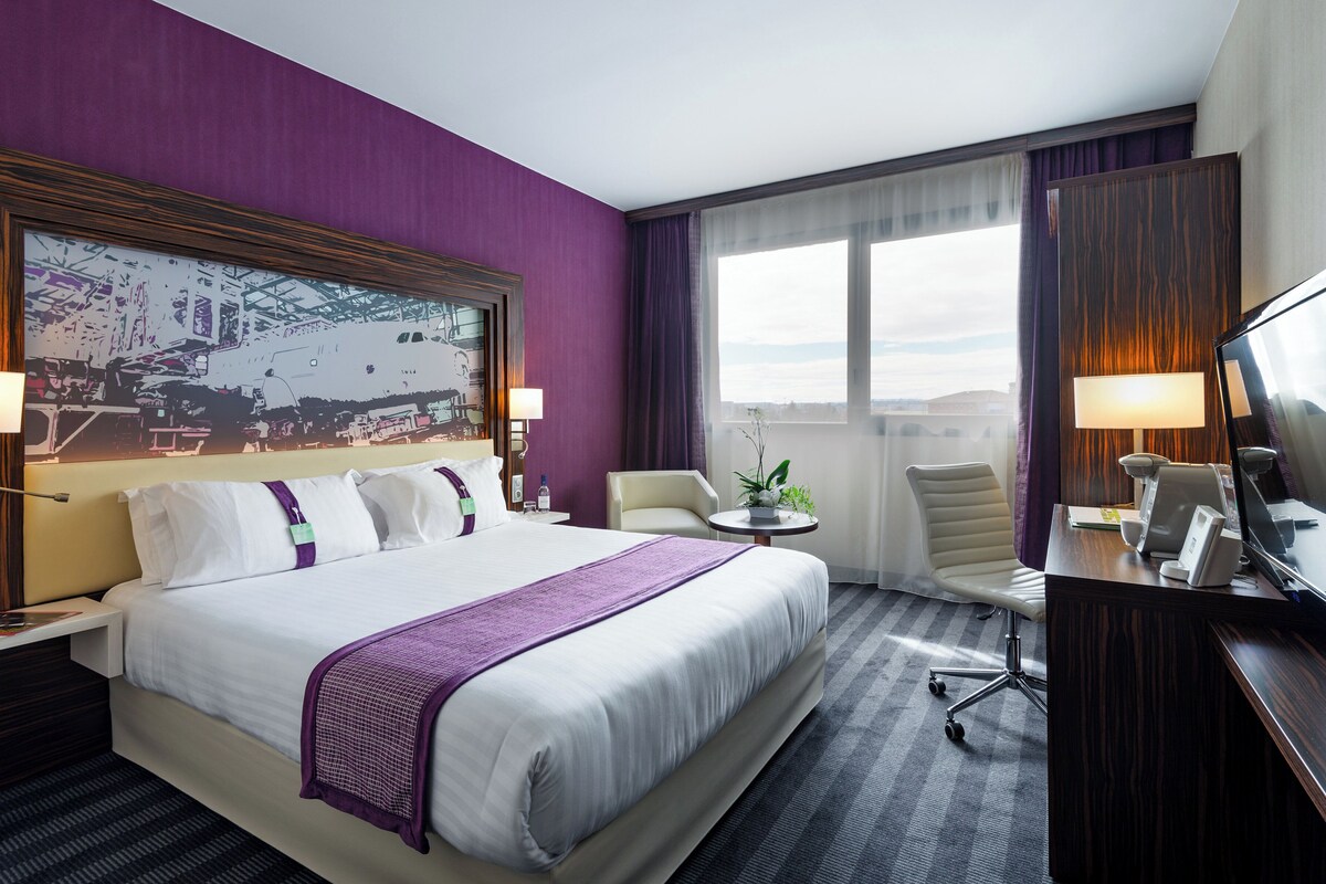 A hotel room with a large bed, a desk with a chair, a TV, and a window with curtains.