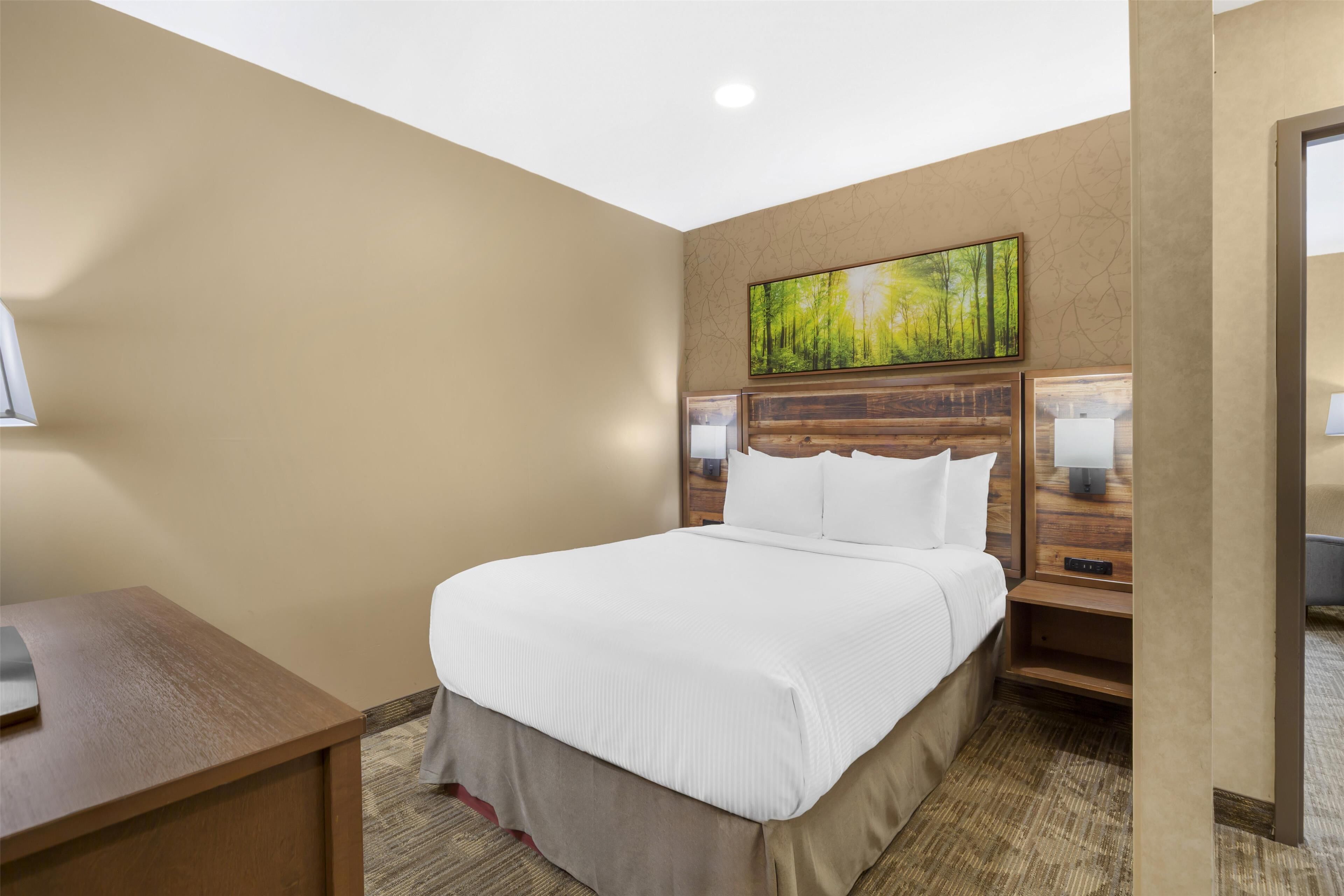 A hotel room with a large bed, a wooden headboard, a painting above the bed, a nightstand with a lamp, and a desk with a lamp.