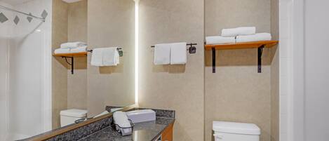 Un baño moderno con encimera de granito, un lavabo blanco, un inodoro y una ducha. Hay dos toalleros con toallas dobladas.