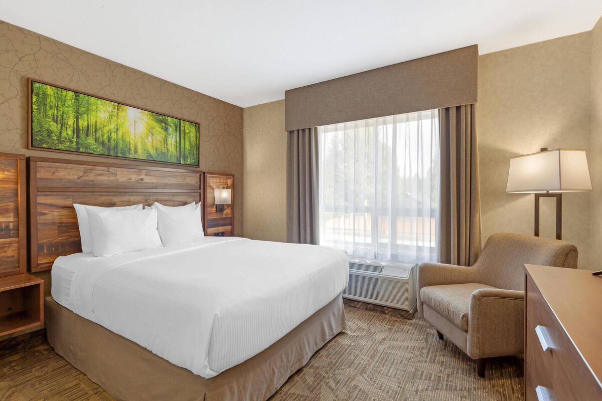 A hotel room with a large bed, a desk, a chair, a window with curtains, and a painting on the wall.