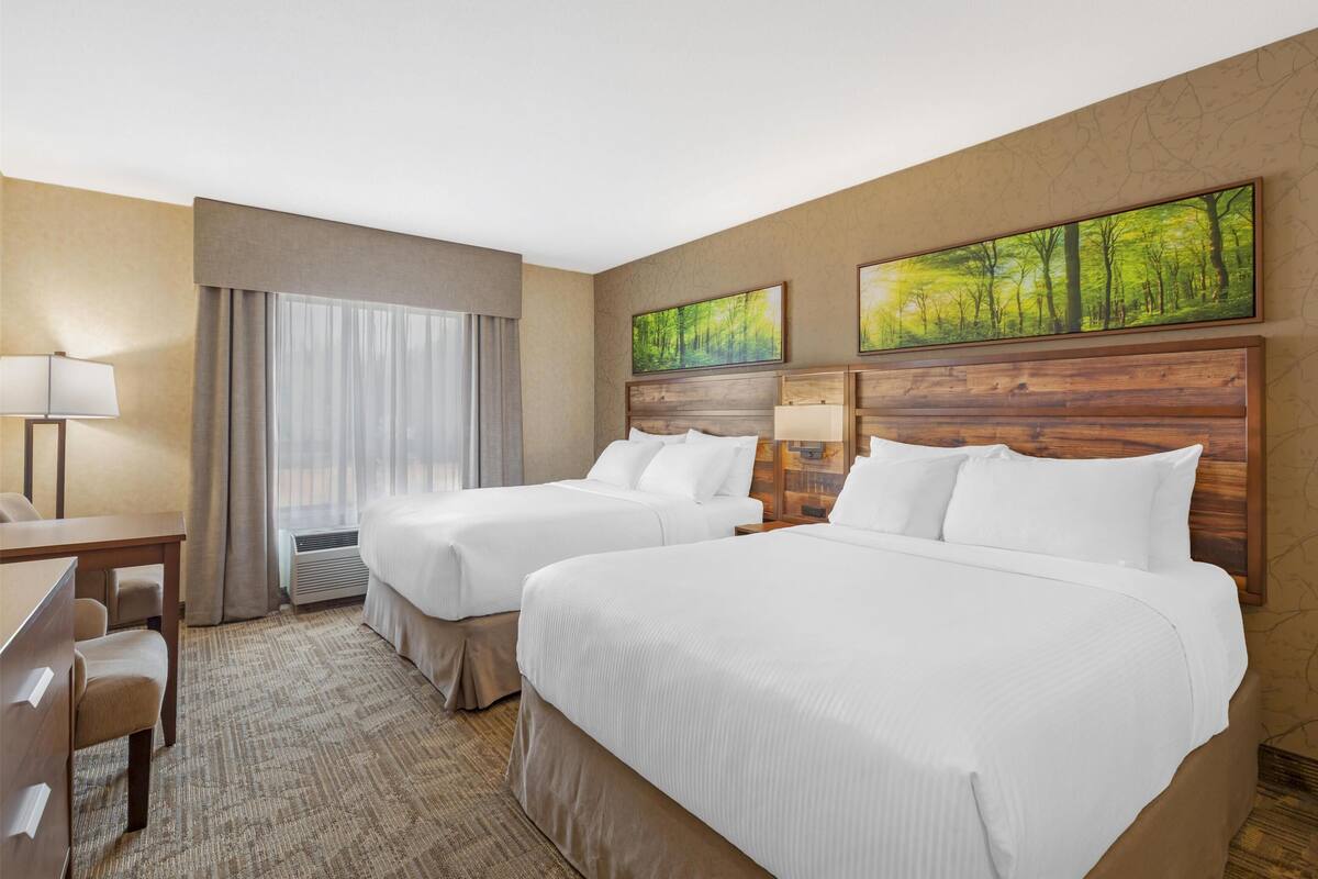 A hotel room with two beds, a desk, and a chair. There are two paintings on the wall and a window with curtains.