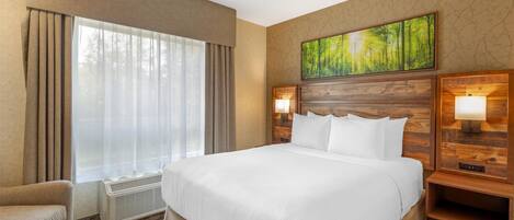 Habitación de hotel con una cama grande, una silla, un ventanal con cortinas, un cuadro en la pared y dos lámparas de noche.