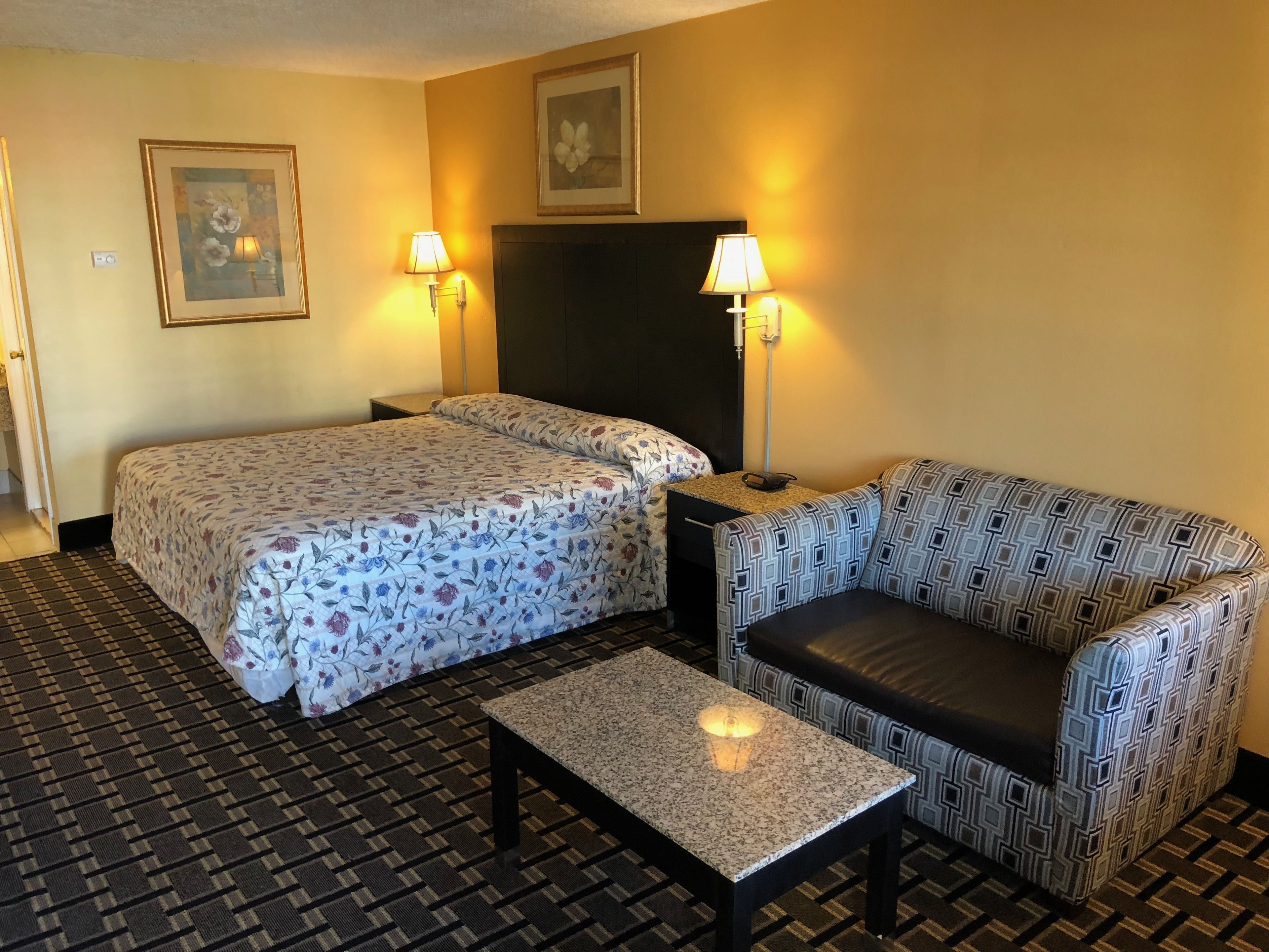 Hotellihuone, jossa on sänky, sohva, pieni pöytä, kaksi seinävalaisinta ja seinällä oleva kehystetty kuva.