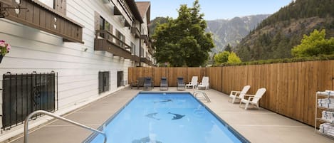 A swimming pool with lounge chairs and a table, surrounded by a wooden fence and a building with balconies.