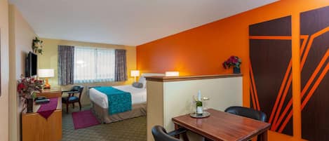 A hotel room with a bed, a desk with a computer, a dining table with chairs, and a large orange accent wall.