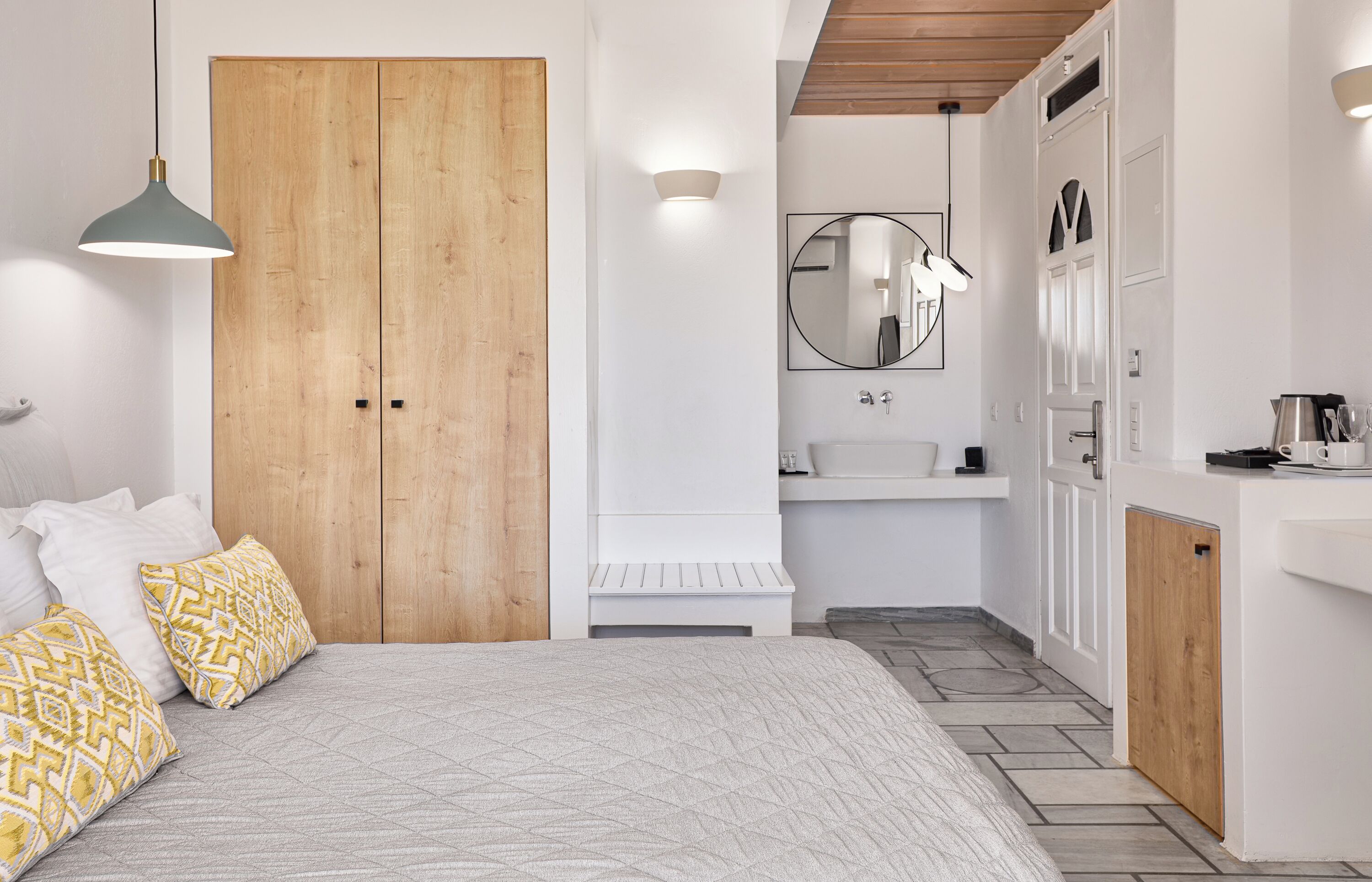 A bedroom with a wooden wardrobe, a bedside table, a bed with patterned pillows, a hanging light, and a small white bench.