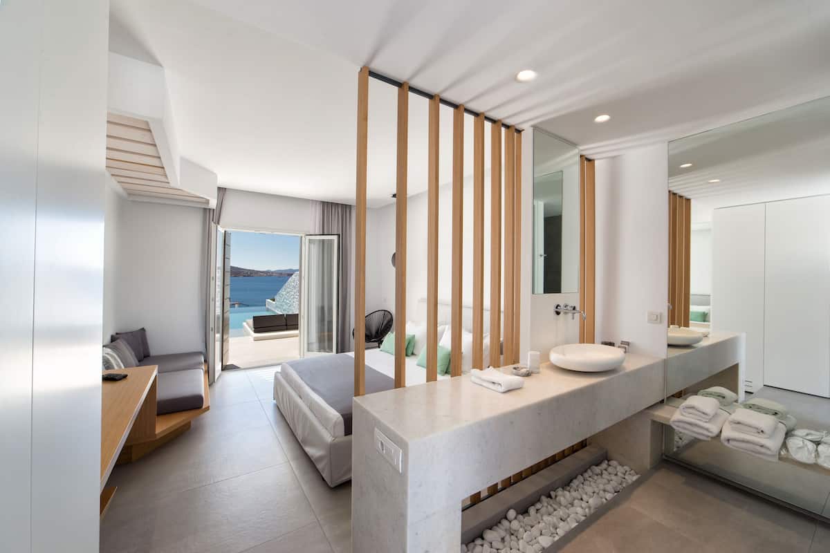 A modern hotel room with a large bed, a sitting area, a bathroom with a large mirror, and a view of the sea.