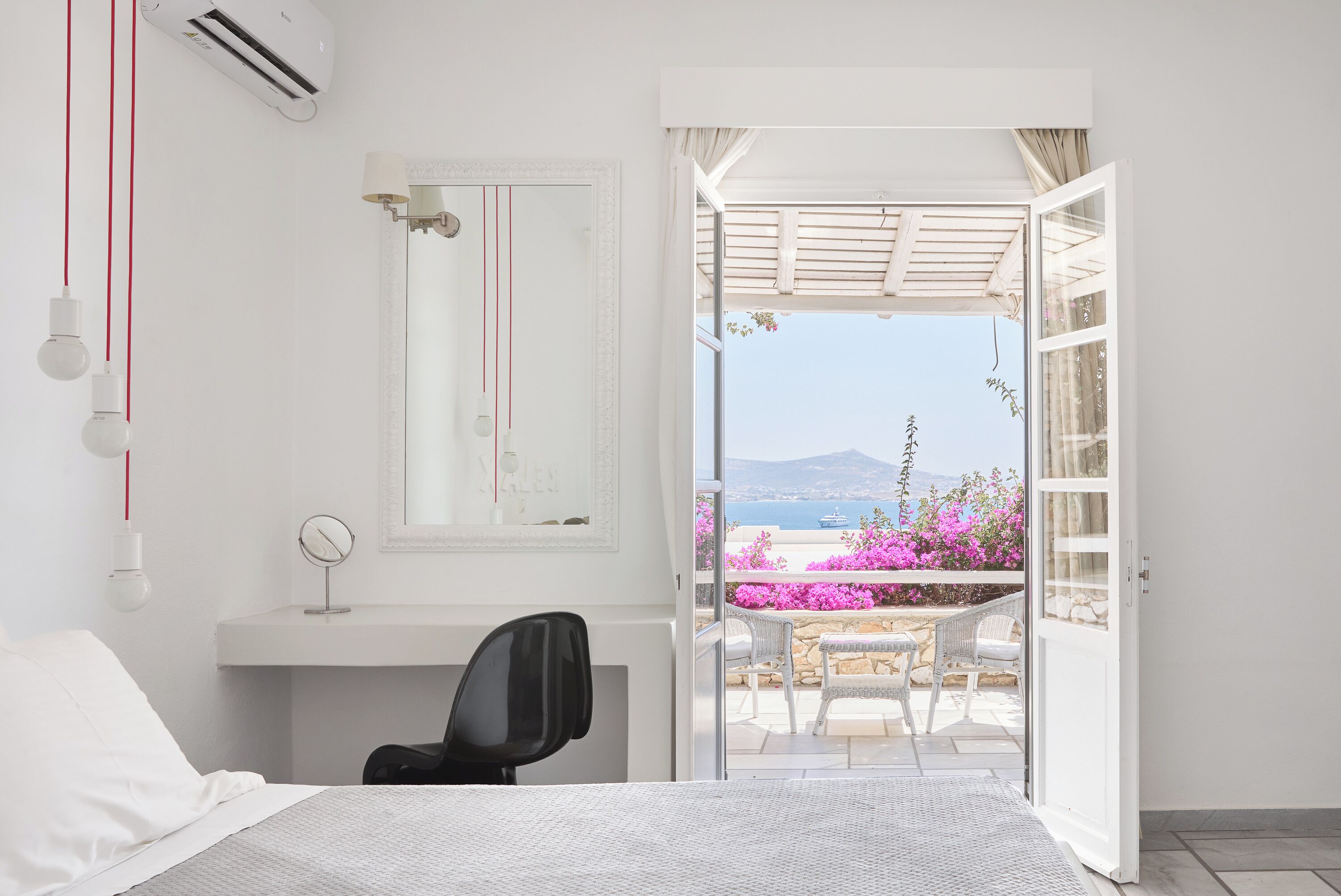 Un dormitorio con cama, escritorio, silla, espejo y puerta abierta que da a un balcón con vistas al mar y flores.