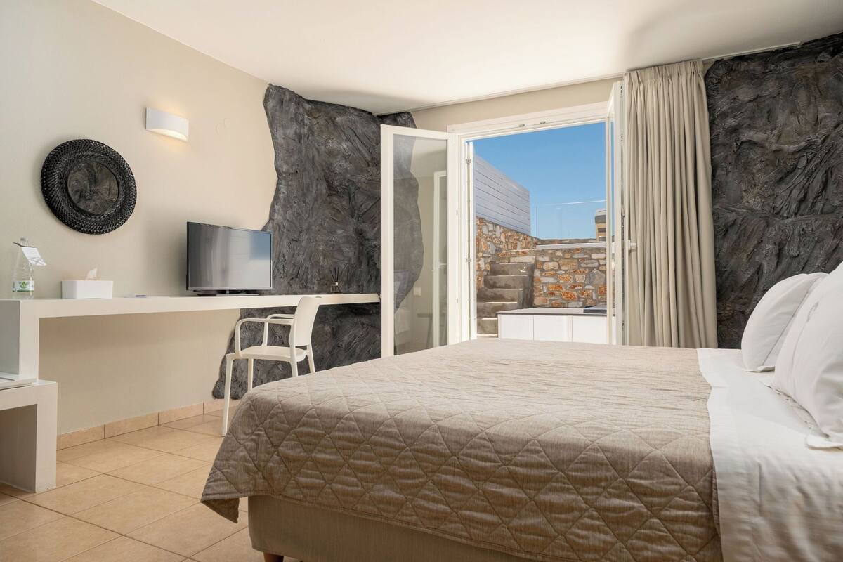 A bedroom with a bed, a desk, a television, and a view of the outside.