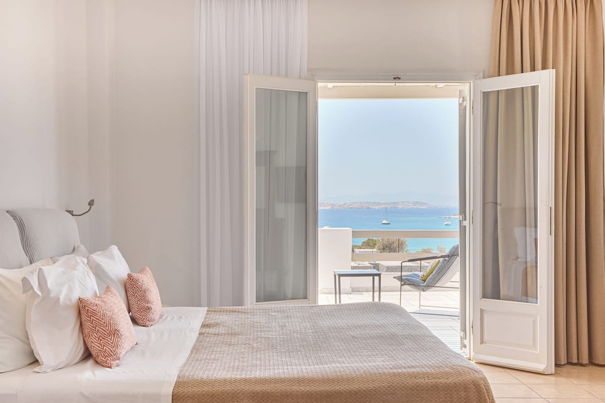 A bedroom with a bed, white curtains, and a view of the sea through an open door.