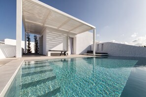 Un área de piscina moderna con una piscina cristalina, una zona de estar cubierta y una pared exterior blanca.
