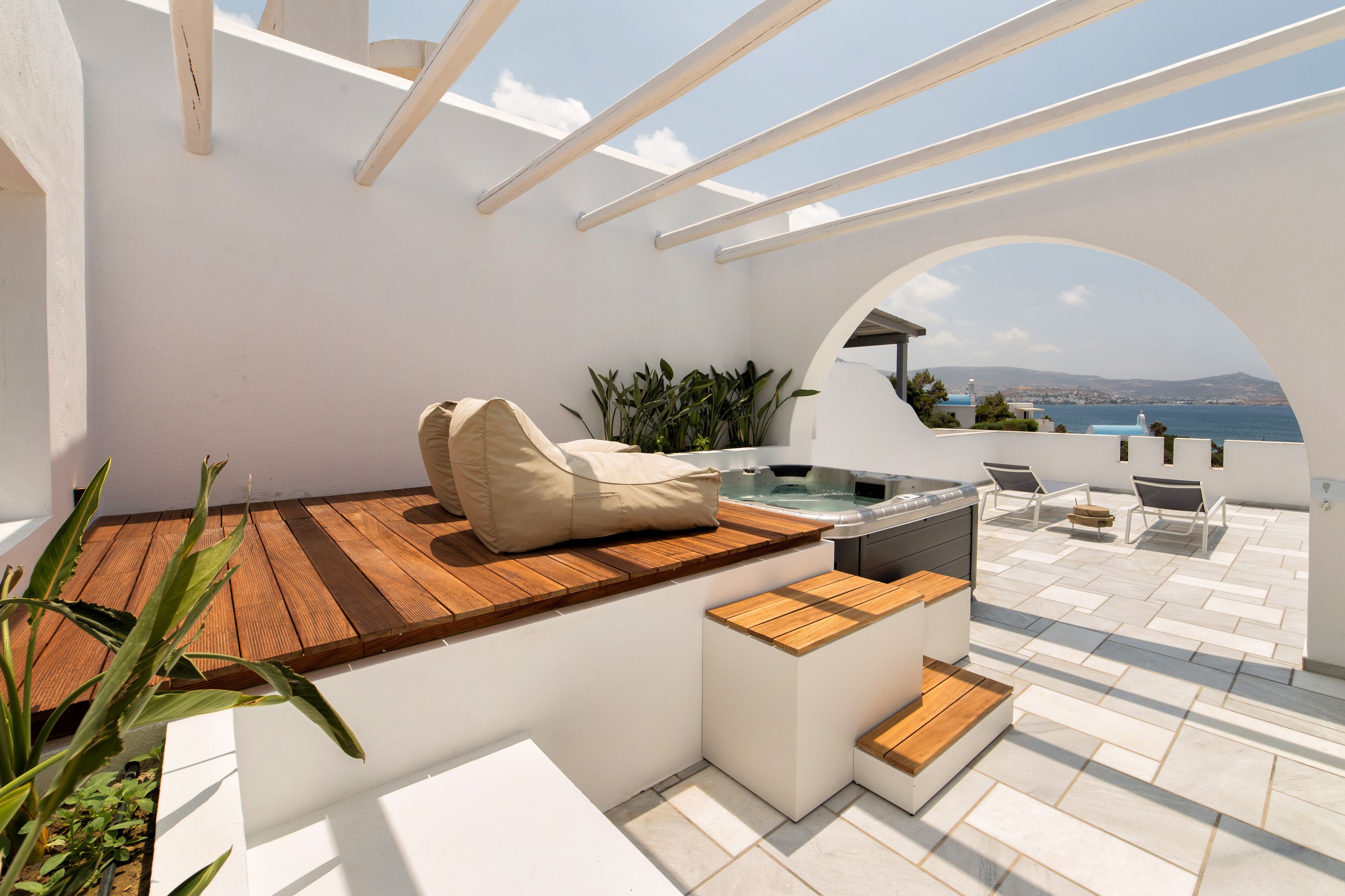 Un patio exterior iluminado por el sol, con una bañera de hidromasaje, asientos de madera y vistas al mar.