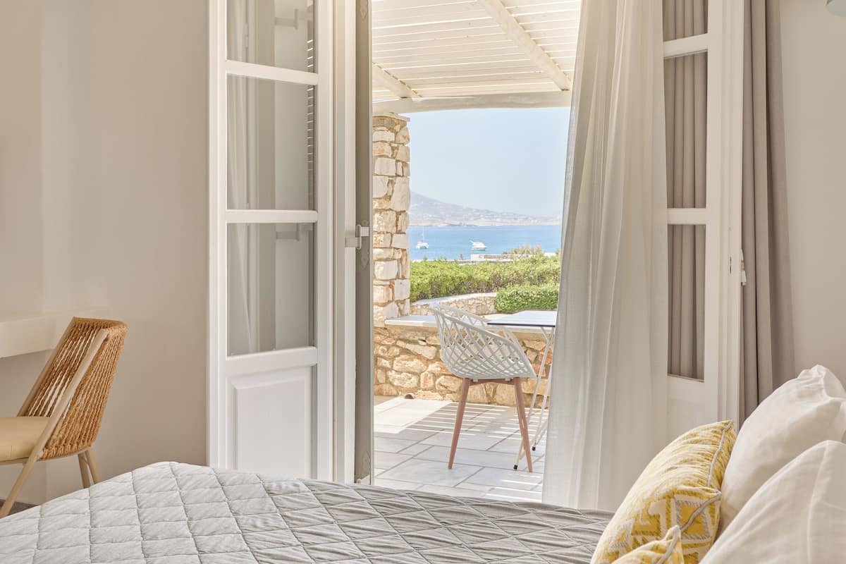 A bedroom with an open door leading to a balcony with a stone wall and a view of the sea.