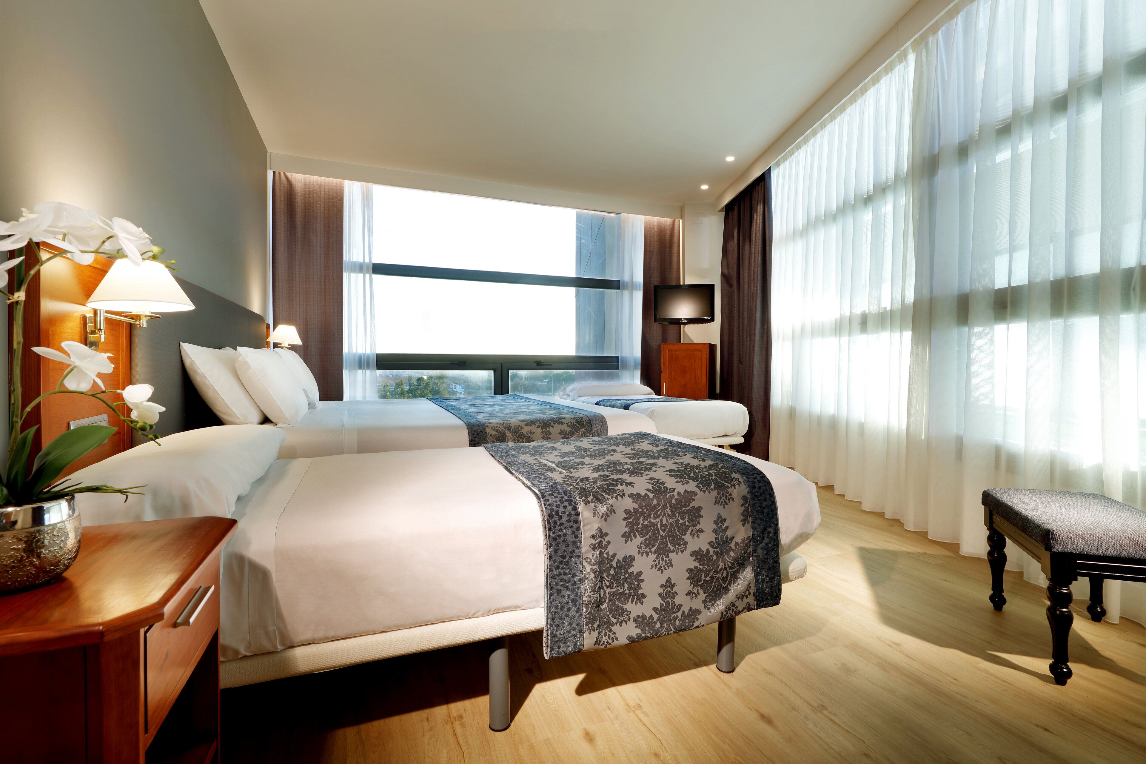 A hotel room with two beds, a bedside table with a lamp, a bench, and a large window with curtains.