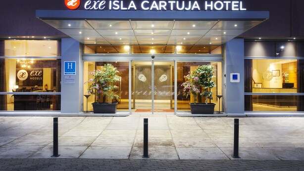 The exterior of the Exe Isla Cartuja Hotel with a glass facade, entrance doors, and potted plants. - Hotel Exe Isla Cartuja (Seville)