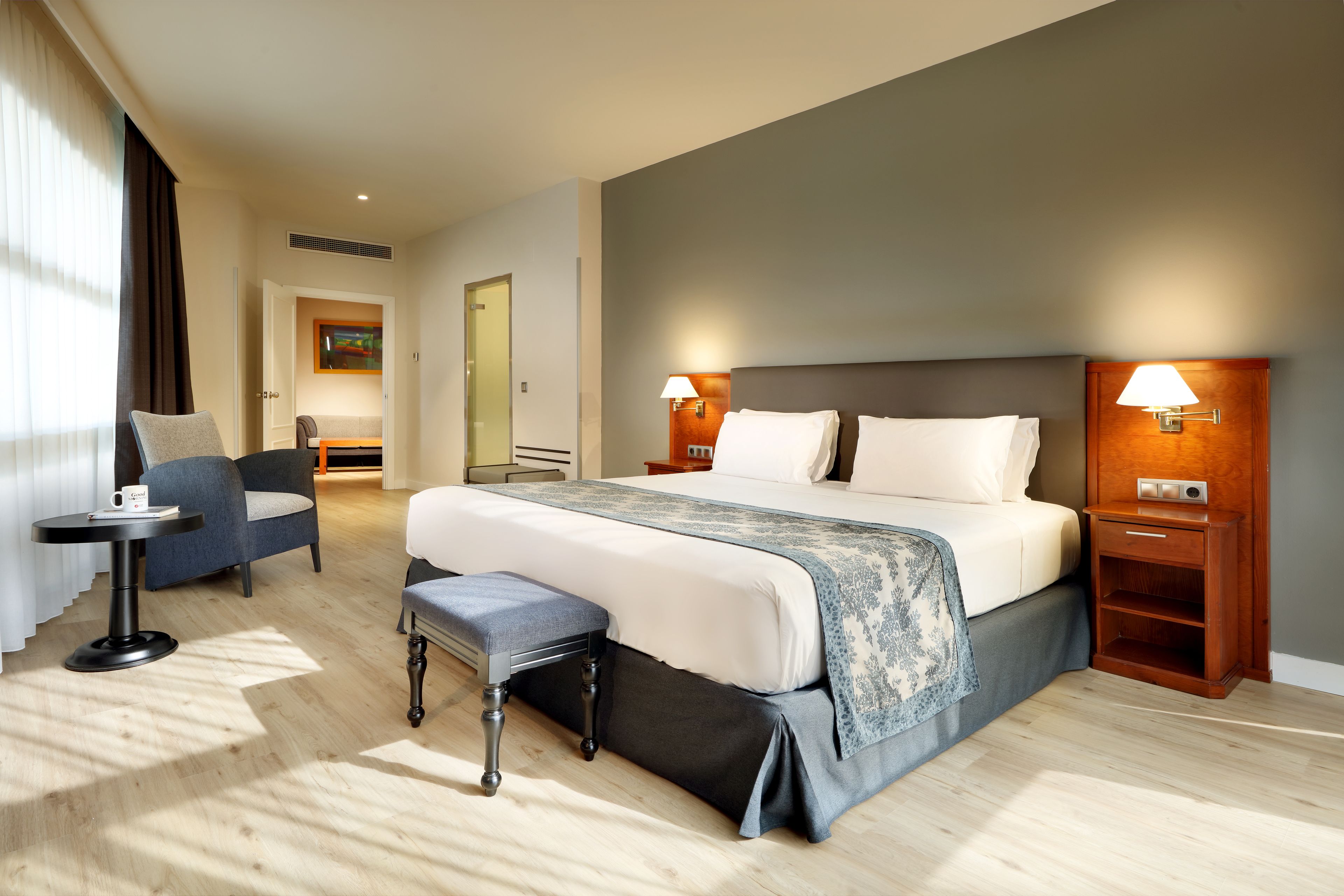 A hotel room with a large bed, two bedside tables with lamps, a small table with a chair, and a bench.