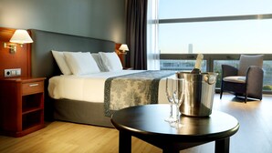 A hotel room with a bed, bedside lamps, a table with a champagne bucket and glasses, a chair, and a view of the city through large windows. - Hotel Exe Isla Cartuja (Seville)