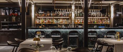 A bar area with multiple shelves of liquor bottles and glasses, a marble counter, and tables with chairs set for dining.