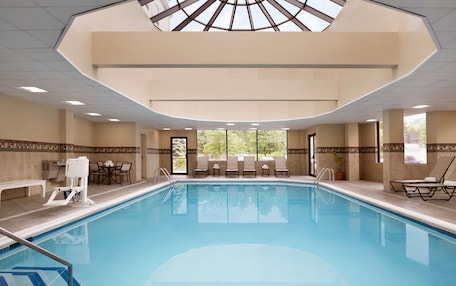 A large indoor swimming pool with a skylight, lounge chairs, and a seating area with tables.