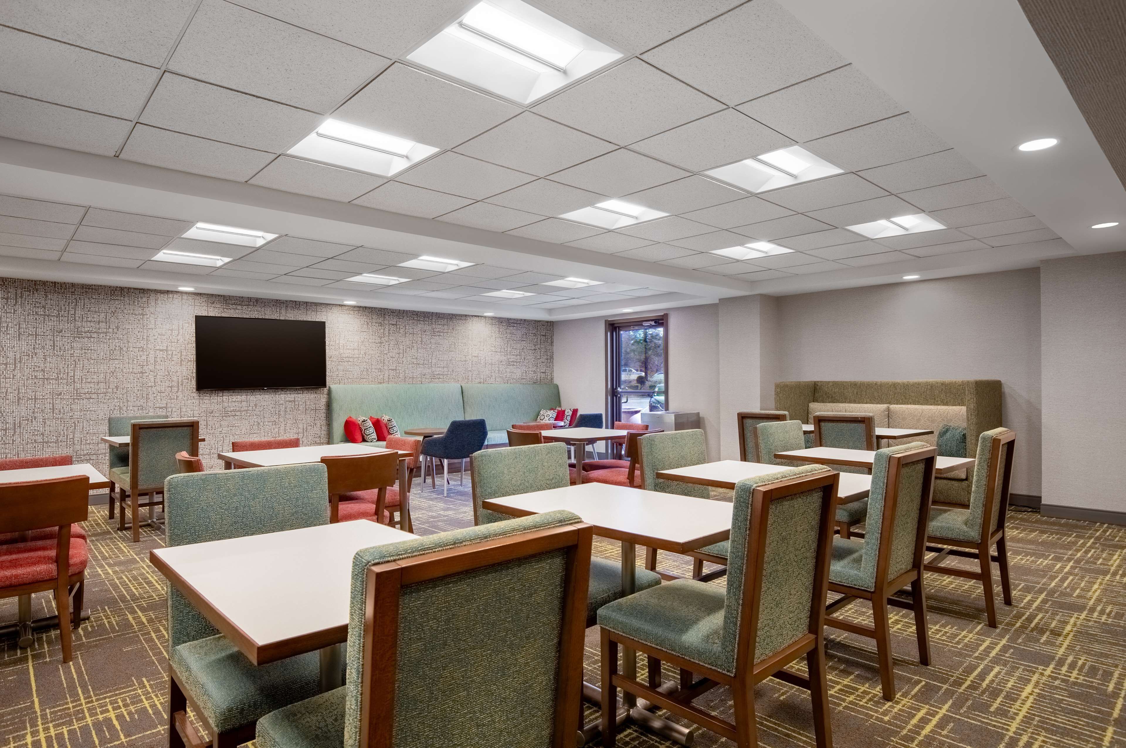 A hotel dining area with tables and chairs, a flat-screen TV on the wall, and a brick-patterned wall.
