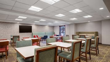 A hotel dining area with tables and chairs, a flat-screen TV on the wall, and a brick-patterned wall.