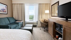 Habitación de hotel con cama, sofá, escritorio con televisor, microondas y vista a zonas verdes.