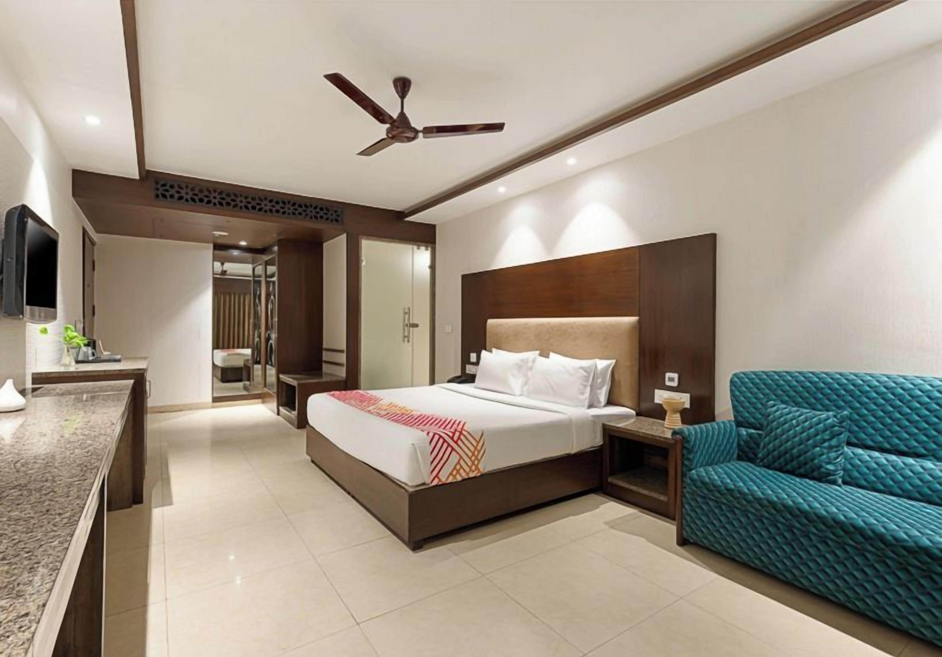 A hotel room with a bed, a desk, a chair, a ceiling fan, and a bathroom in the background.