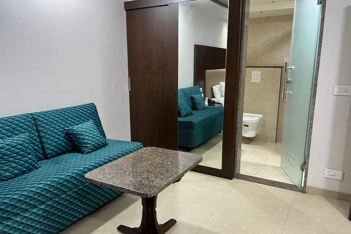 A room with a teal sofa, a small table, and a mirror reflecting a bathroom with a toilet and a bed.
