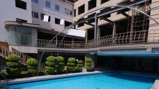A swimming pool area with a covered bridge, a multi-story building, and neatly trimmed bushes. - Hotel Amar (Agra)