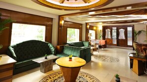 A spacious lobby with a high ceiling, a central seating area with green sofas, a wooden reception desk, and a large decorative ceiling feature. - Hotel Amar (Agra)
