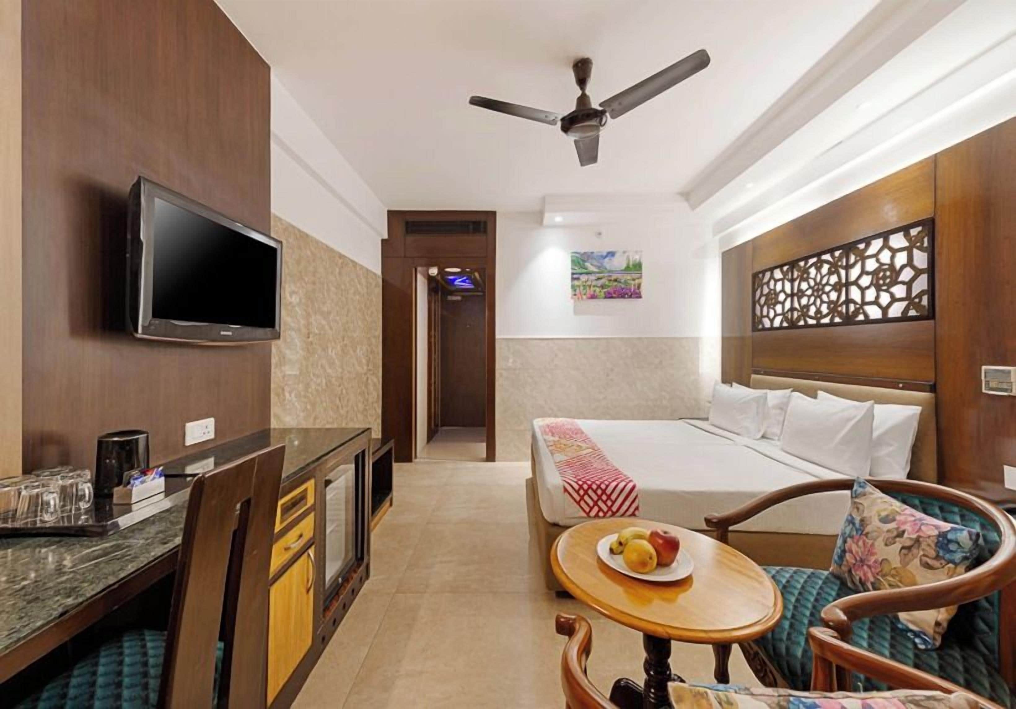 A hotel room with a bed, a desk with a flat-screen TV, a ceiling fan, and a small table with a plate of fruit.