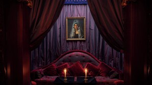 Um quarto escuro com um sofá de veludo vermelho, duas velas sobre uma mesa e um retrato emoldurado na parede.