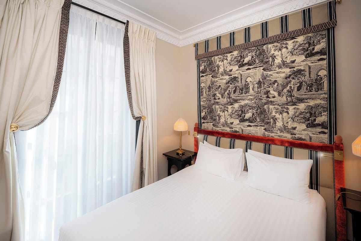 A neatly made bed with white linens, a classic headboard with a detailed design, and a large tapestry depicting a historical scene on the wall.