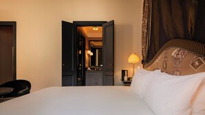 Quarto de hotel com uma cama grande, uma escrivaninha, uma cadeira, uma mesinha de cabeceira, uma luminária e uma porta para o banheiro.