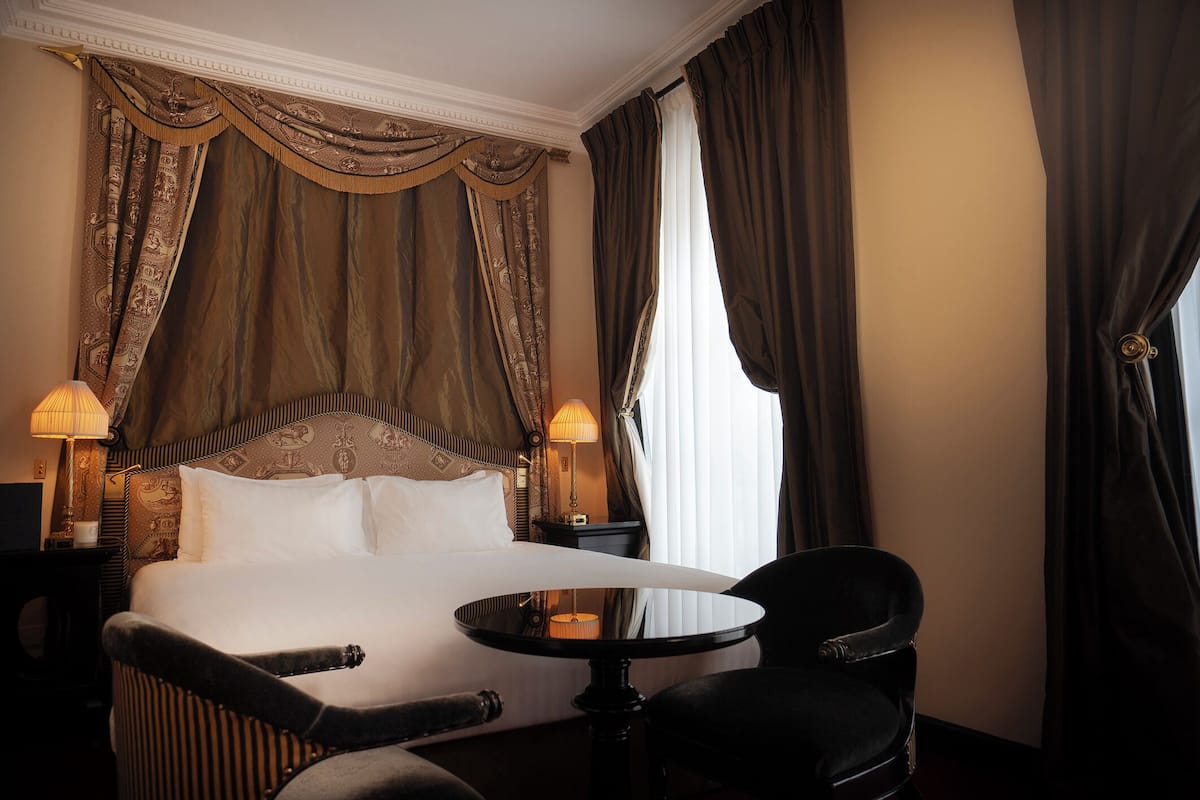 A hotel room with a large bed, two bedside lamps, a round table, and a chair.