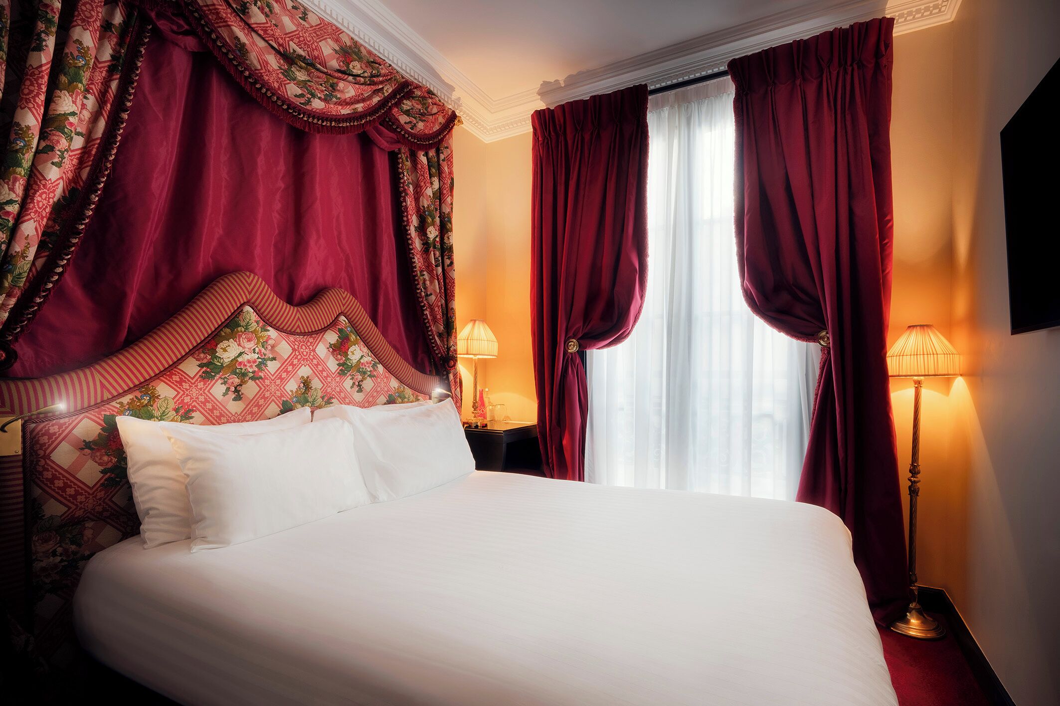A hotel room with a large bed, red curtains, a patterned headboard, and two bedside lamps.