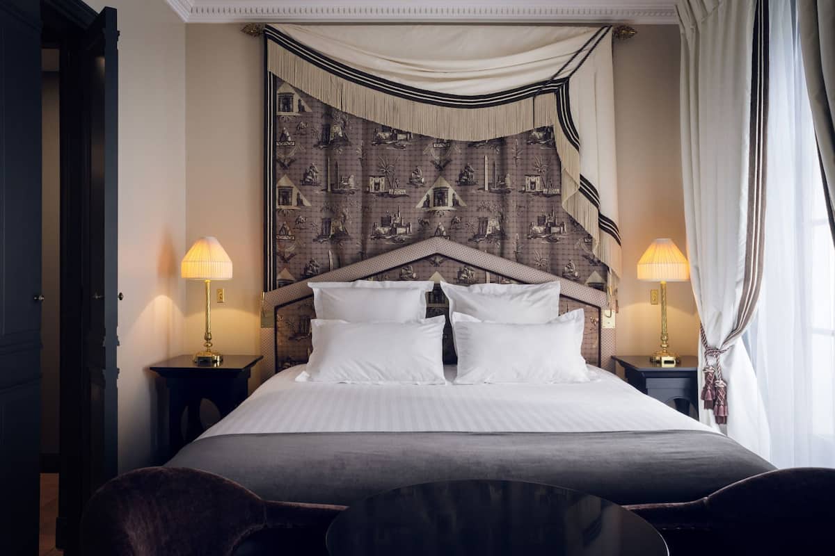 A hotel room with a large bed, two bedside tables with lamps, a headboard with a tapestry, and a door on the left.