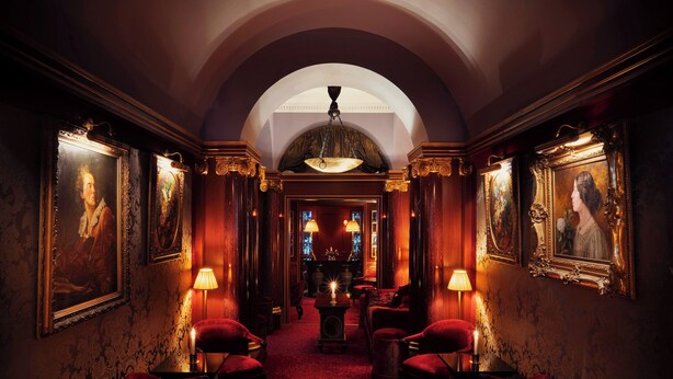 Um corredor com pouca luz, cadeiras de veludo vermelho, decorações elaboradas nas paredes e um candelabro.