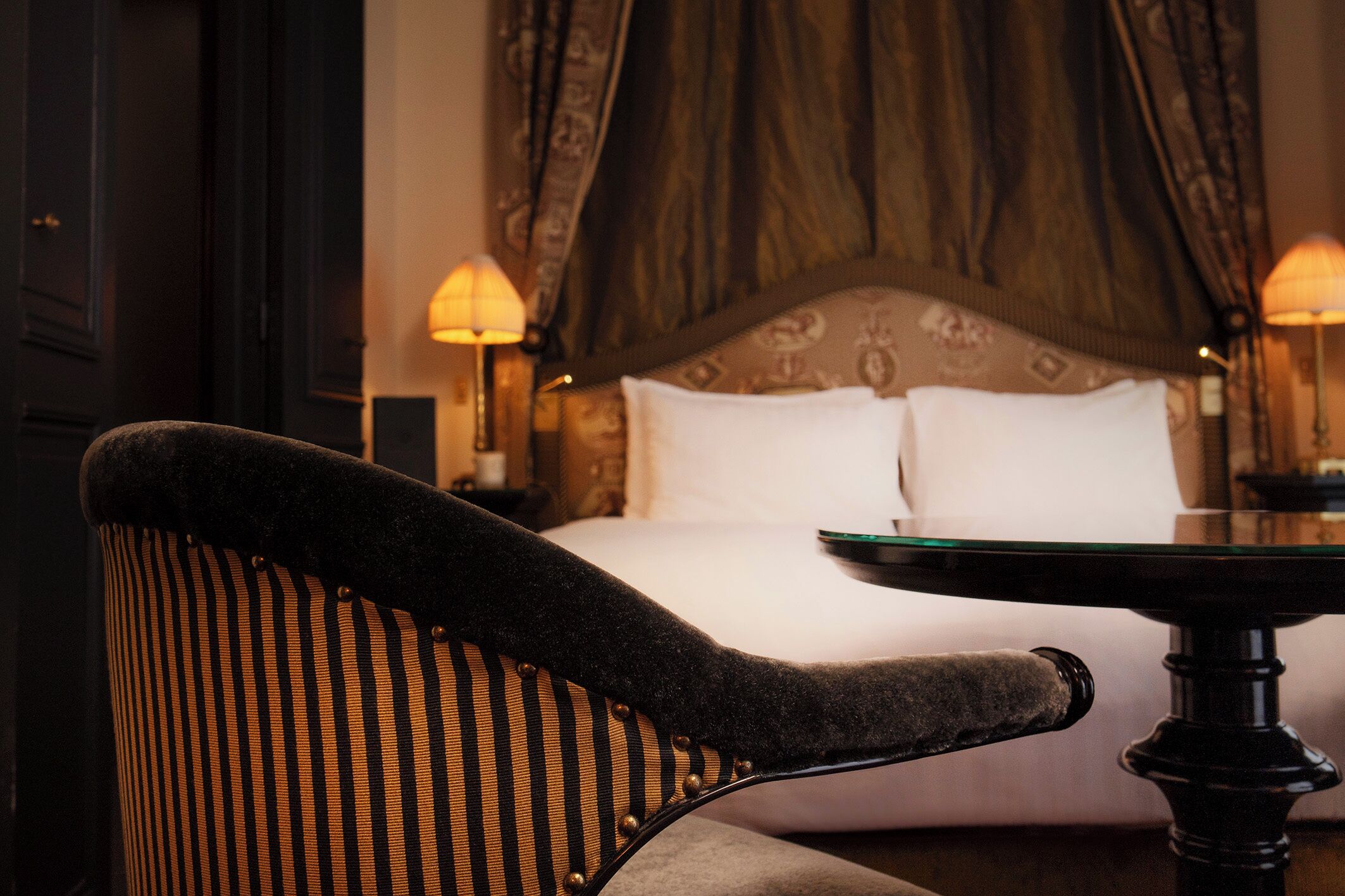 A hotel room with a bed, two bedside lamps, a table, and a chair.